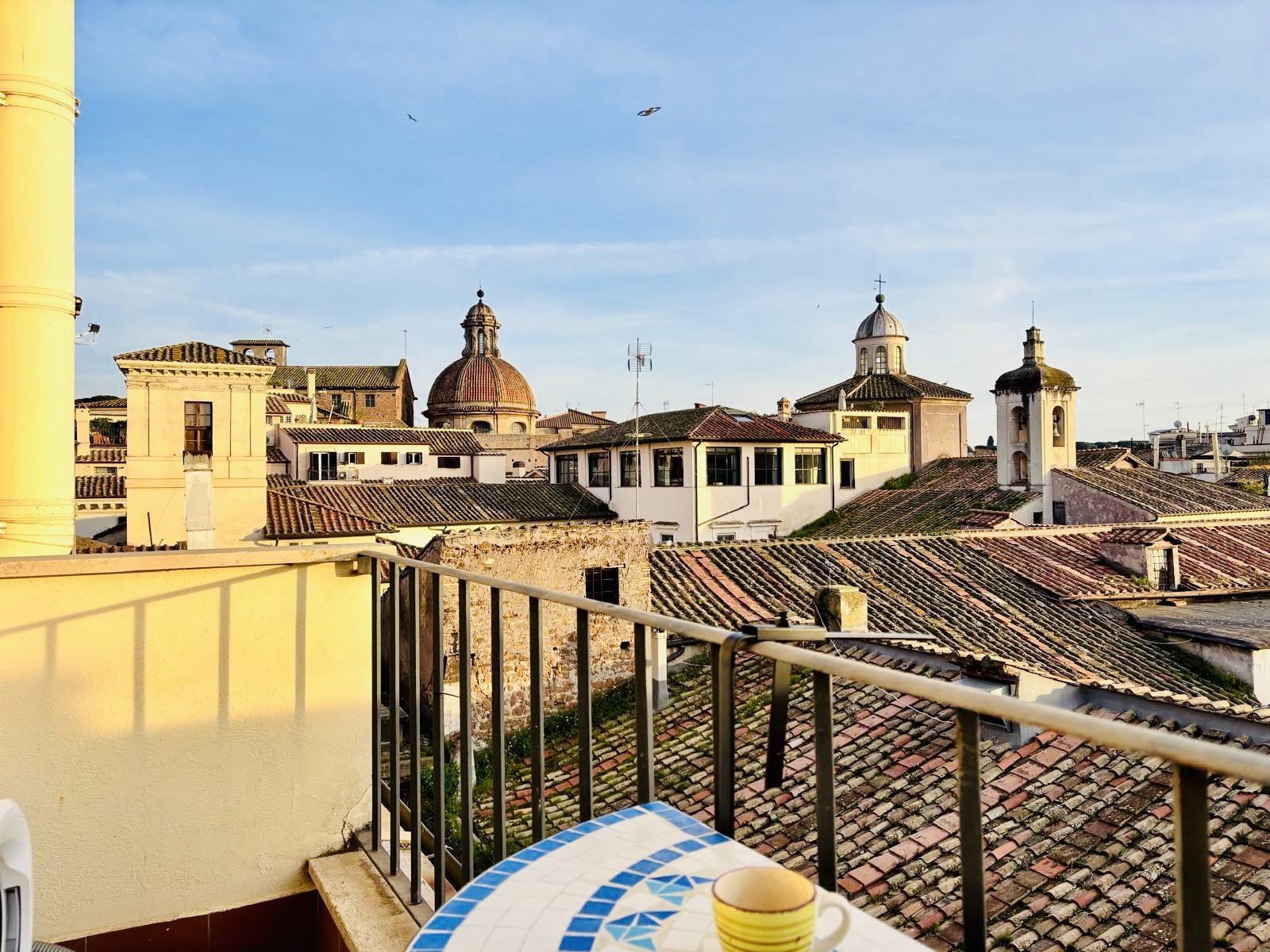 Golden light on the balcony railing, with a coffee cup on the mosaic table and the rooftops and domes of old Rome beyond