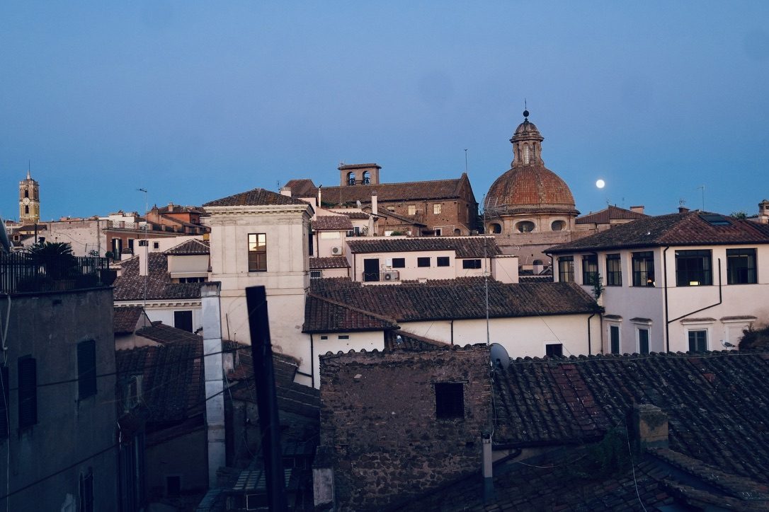 Blue hour over the rooftops, with the moon rising beside a terracotta dome